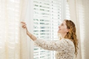 womain opening white curtains to reveal window with blinds